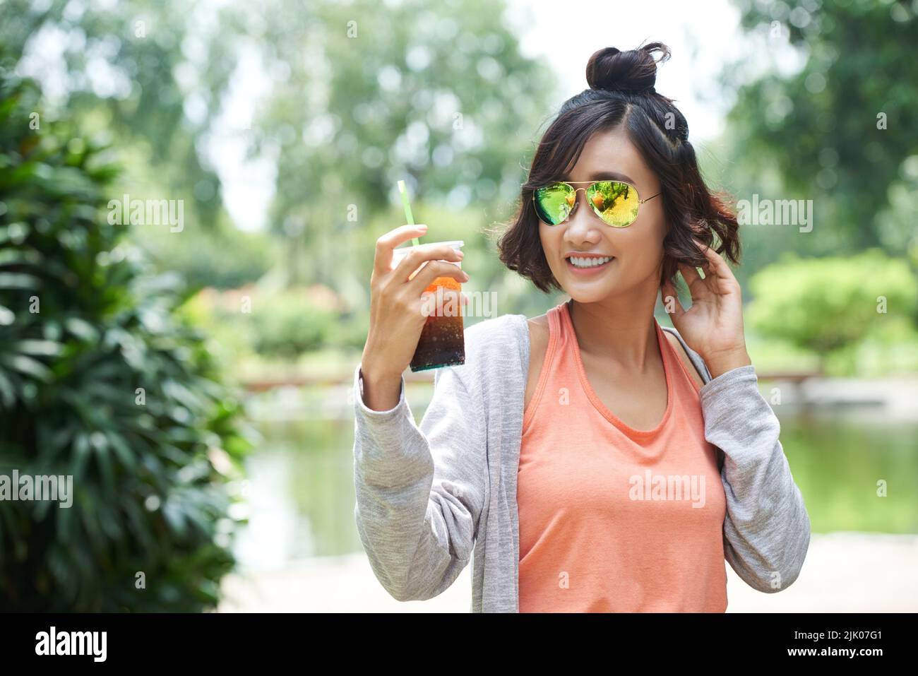 Portrait à la taille haute d'une femme asiatique gaie portant des lunettes de soleil en regardant loin avec un sourire éclatant tout en tenant une tasse de thé glacé à la main, parc public vert sur l'arrière-plan Banque D'Images
