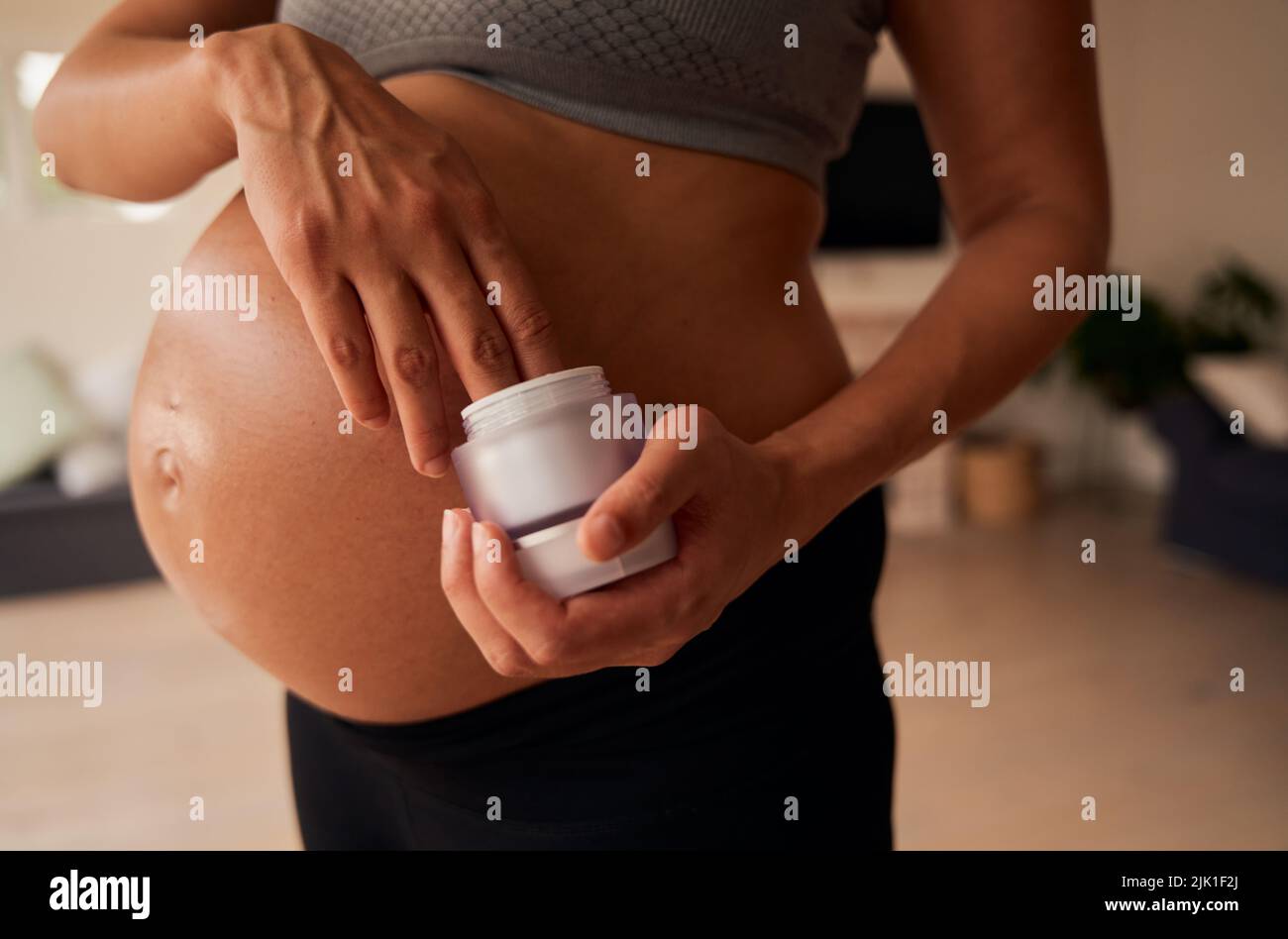 Torse de femme caucasienne enceinte debout, appliquant de la crème hydratante sur son ventre. Soins de la peau. Un style de vie actif dans une maison moderne en journée. Banque D'Images