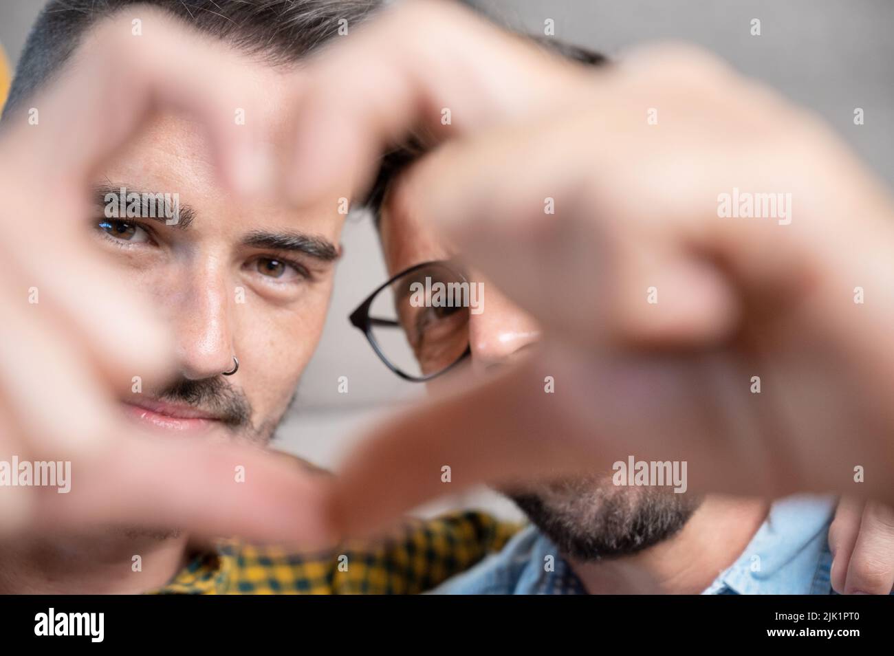 Jeune couple gay souriant confiant faisant le symbole de coeur avec les mains à la maison. Photographie de haute qualité. Banque D'Images