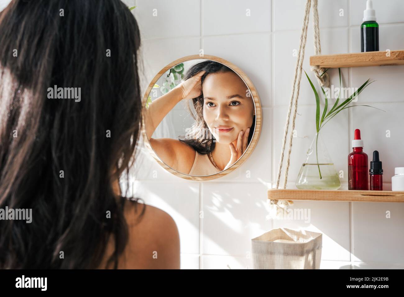 Belle femme hispanique regardant dans le miroir dans la salle de bains écologique blanche. Shleves en bois et bouteilles de cosmétiques réutilisables. Concept de wellnes Banque D'Images