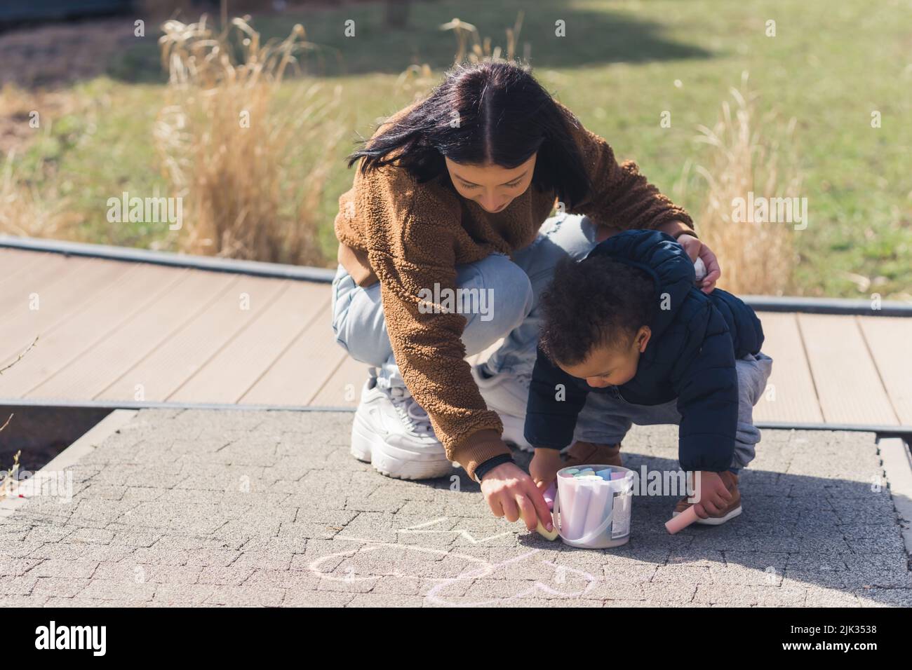 La mère caucasienne et son petit enfant afro-américain portant des vêtements d'hiver et dessinant des coeurs sur le trottoir à l'aide de crayons colorés - regard de fond de parc d'en haut. Photo de haute qualité Banque D'Images