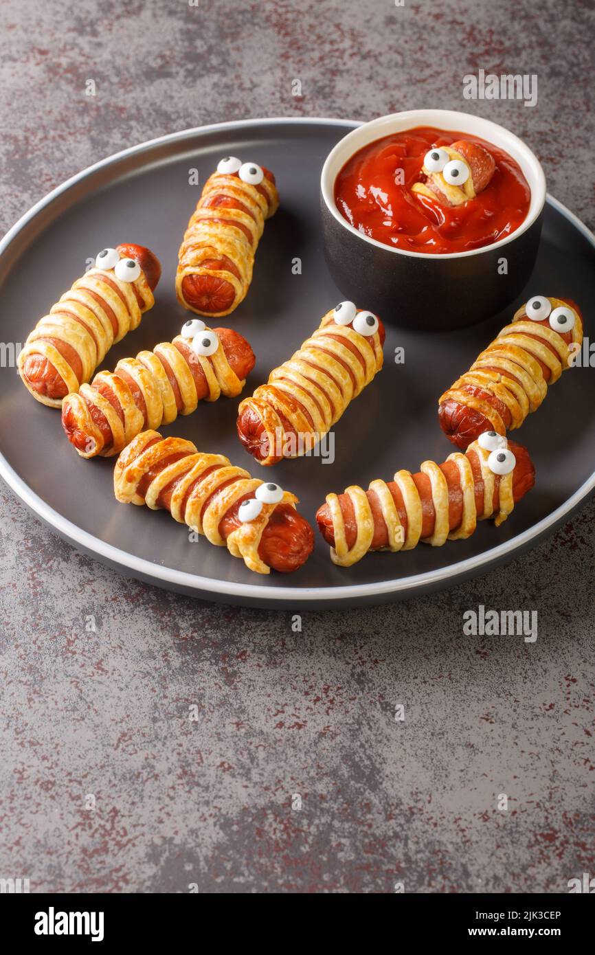 Des saucisses de momie effrayantes dans la pâte avec les yeux drôles sur la table de gros plan. Décorations amusantes. Nourriture pour Halloween. Verticale Banque D'Images