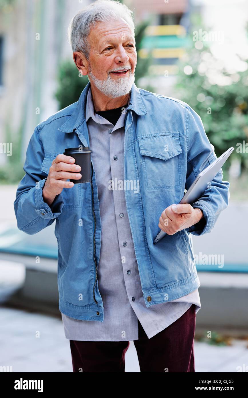 Un étudiant senior heureux est debout dans la rue et tient son ordinateur portable et une tasse de café. Étudiant senior dans la rue se précipitant à une conférence. Banque D'Images