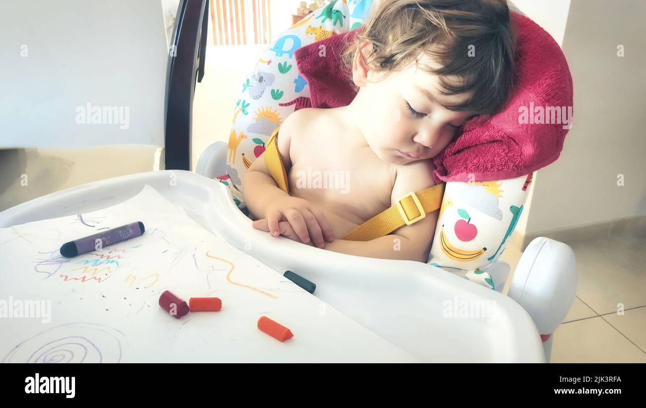 Un petit garçon s'est endormi dans sa chaise haute après avoir joué avec des crayons de couleur Banque D'Images