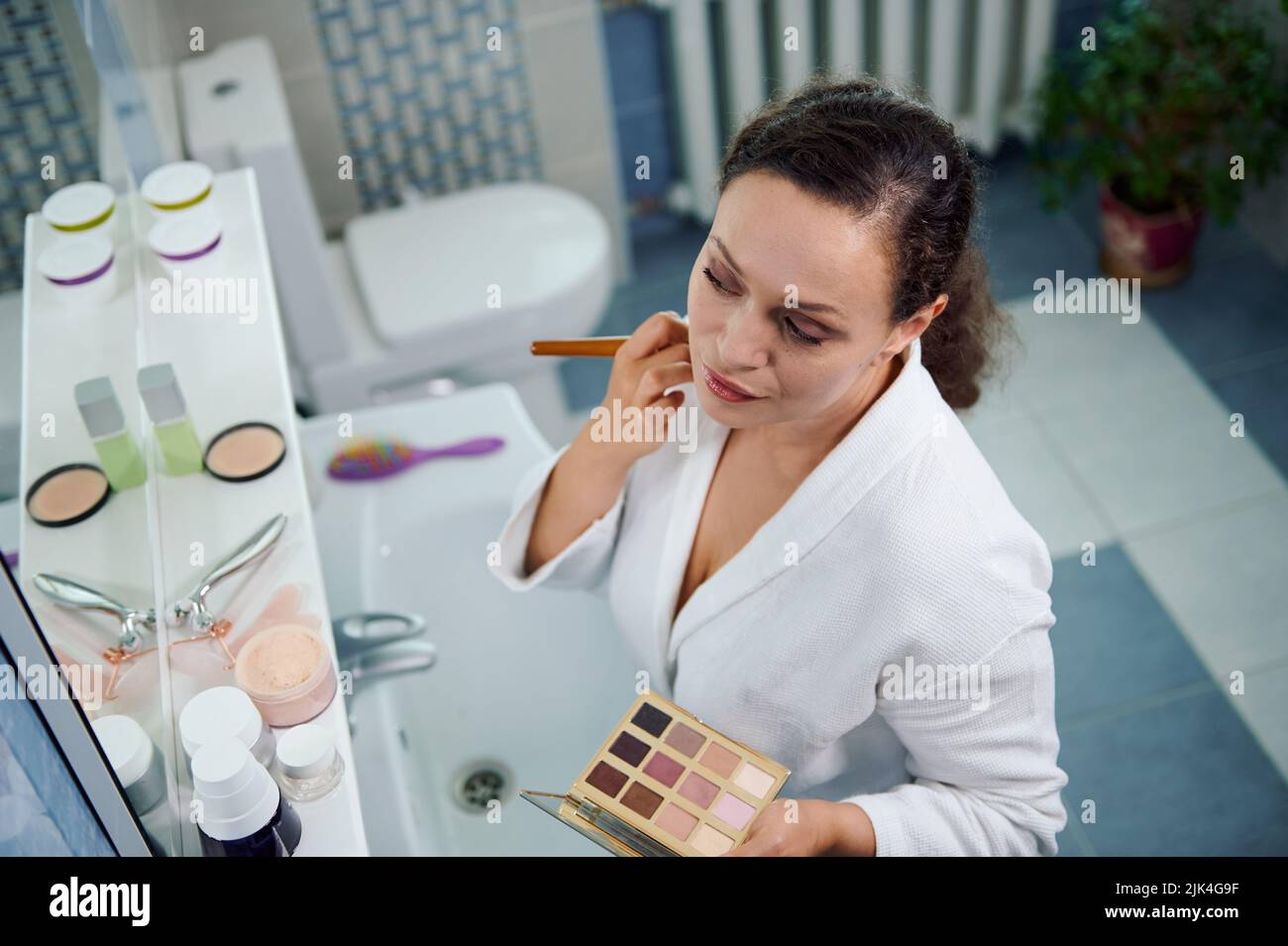 Femme étourdissante applique de la poudre matifiante sur son visage, satisfait de son apparence, regarde son reflet dans le miroir Banque D'Images
