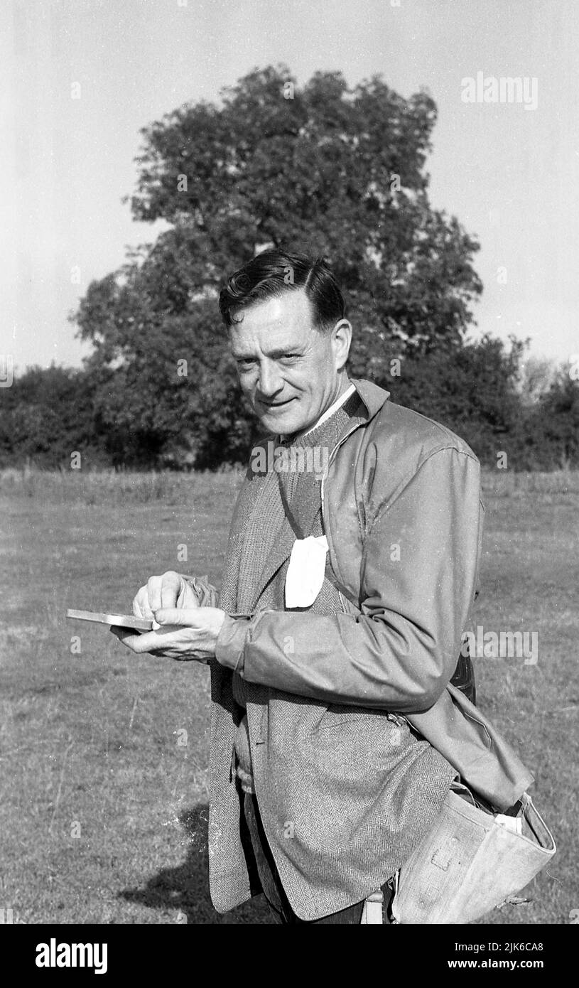 1960s, historique, en plein air, un pêcheur grossier, tenant un petit  plateau à la recherche d'un crochet, Angleterre, Royaume-Uni. Vêtements de  pêche de l'époque, sac à bandoulière en toile, veste imperméable et