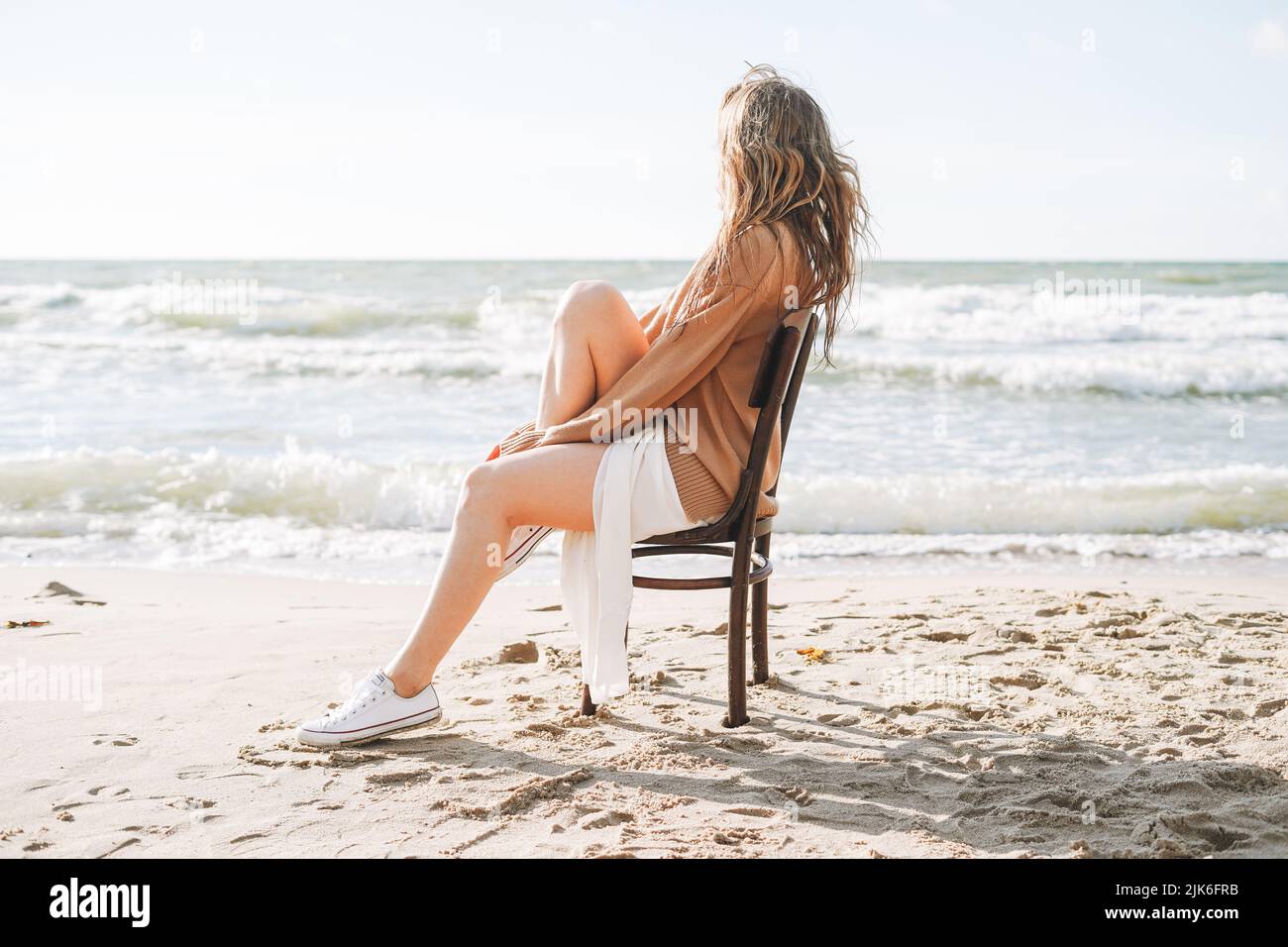 Jeune femme insouciante belle avec de longs cheveux dans le chandail assis sur une chaise sur la plage de mer Banque D'Images