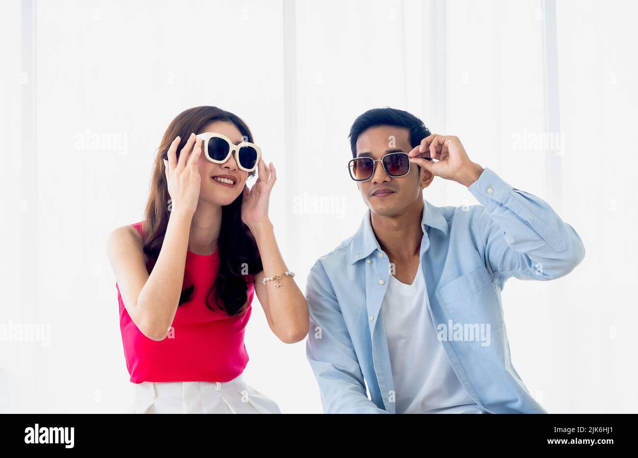 Joyeux couple asiatique en été, jeune homme et femme portant des lunettes de soleil avec le sourire, assis dans la chambre lumineuse de l'hôtel près du rideau blanc et du verre Banque D'Images