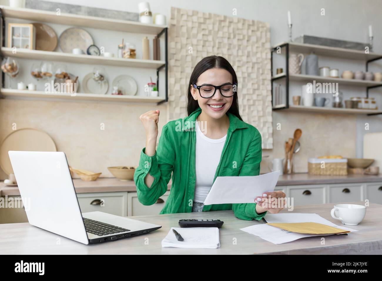 Une femme heureuse en lunettes et une chemise verte à la maison faisant du travail en papier a obtenu de bons résultats, regardant des documents et heureux, tenant main vers le haut geste de la réussite et de la victoire Banque D'Images