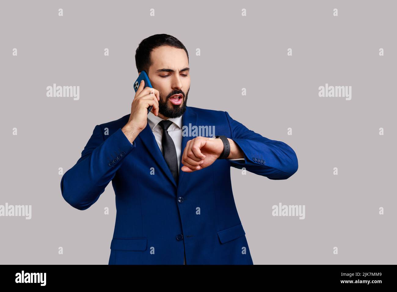 Portrait de beau barbu homme parlant téléphone et regardant montre-bracelet, prendre rendez-vous, attendre la date, porter un costume officiel. Prise de vue en studio isolée sur fond gris. Banque D'Images