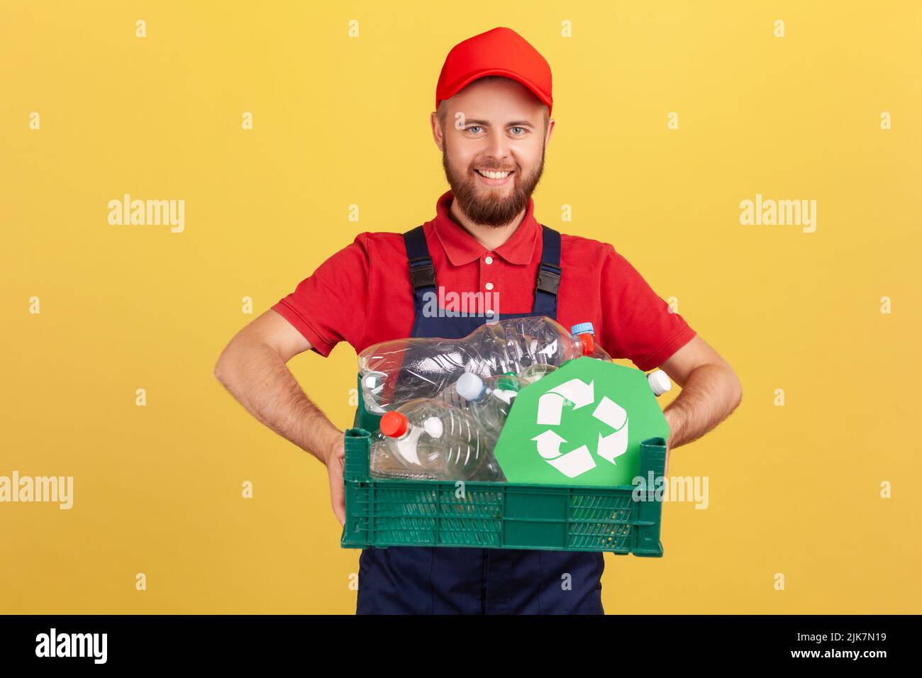 Portrait d'un travailleur à barbe heureux debout et tenant une boîte avec des bouteilles en plastique et une affiche de recyclage, portant une combinaison et une casquette rouge. Studio d'intérieur isolé sur fond jaune. Banque D'Images