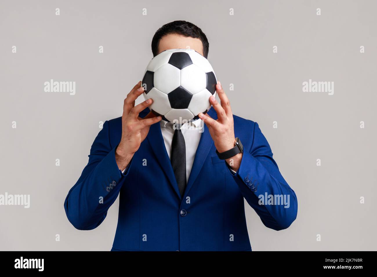 Portrait d'un homme d'affaires anonyme inconnu qui soutient son équipe préférée en couvrant le visage avec un ballon de football, portant un costume de style officiel. Prise de vue en studio isolée sur fond gris. Banque D'Images