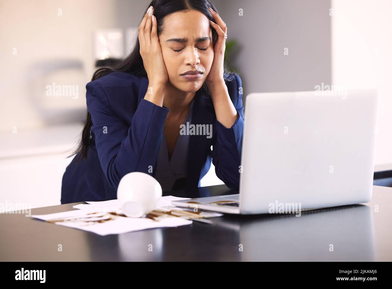 Cette journée peut encore s'aggraver. Une jeune femme d'affaires qui cherche contrariée de renverser du café sur un ordinateur portable et de la paperasse sur une table. Banque D'Images