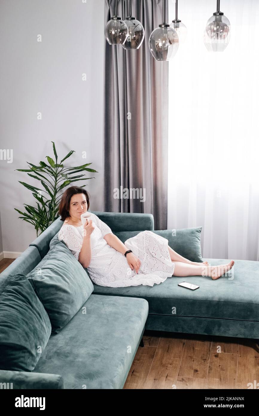 Une femme boit une tasse de café dans une atmosphère chaleureuse le matin. Vraie femme d'âge moyen plus grande en robe blanche se relaxant sur le canapé, moderne Banque D'Images