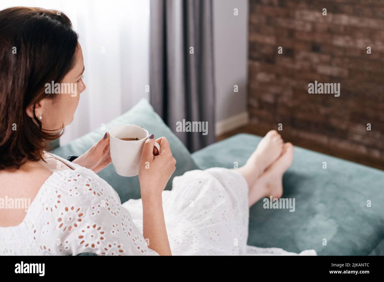 Une femme boit une tasse de café dans une atmosphère chaleureuse le matin. Vraie femme d'âge moyen plus grande en robe blanche se relaxant sur le canapé, moderne Banque D'Images