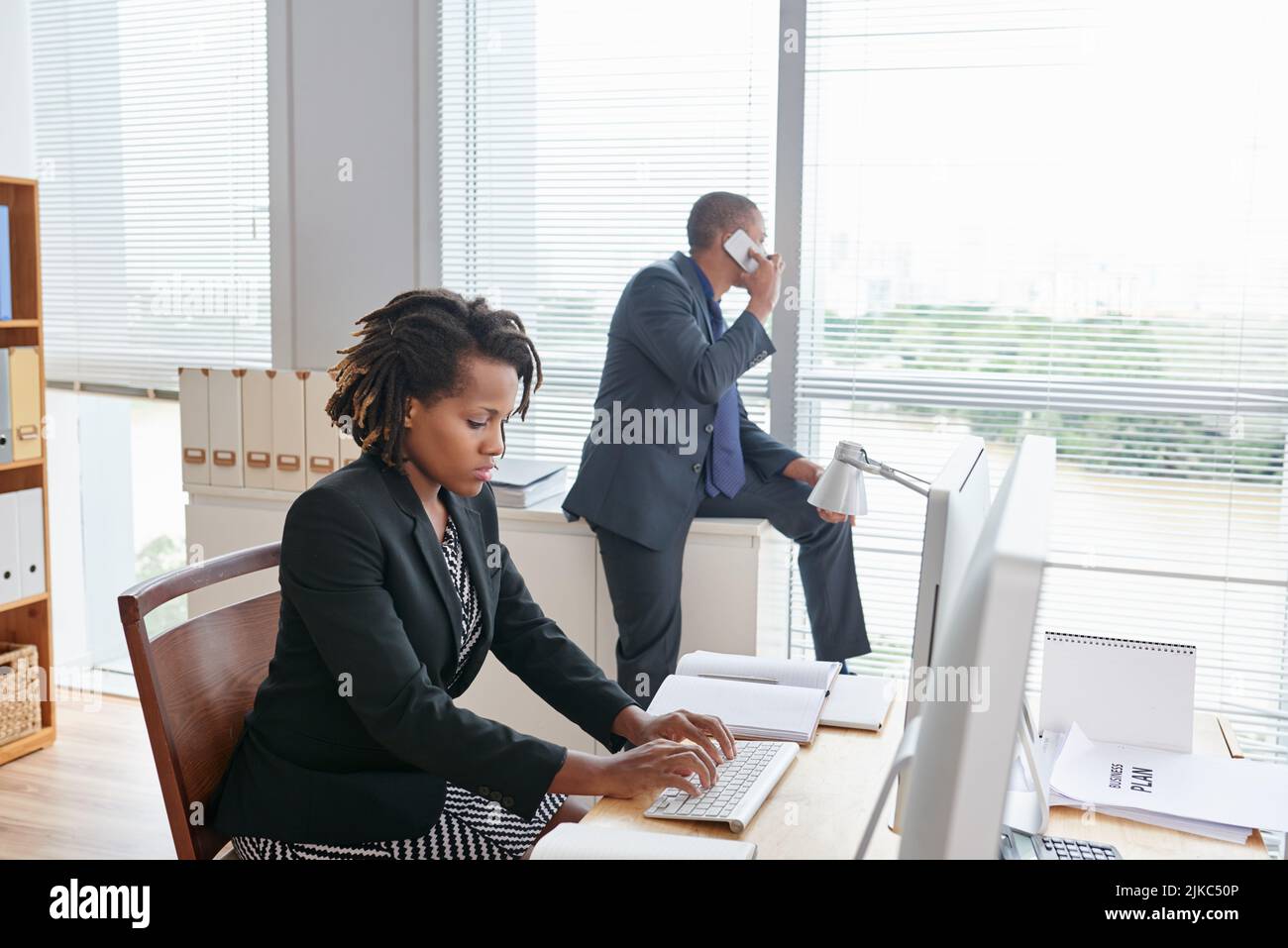 Un bureau ouvert très occupé : une jolie jeune employée de col blanc est assise devant l'ordinateur et travaille sur un projet prometteur tandis que son collègue de sexe masculin discute des détails de la commande avec le client Banque D'Images