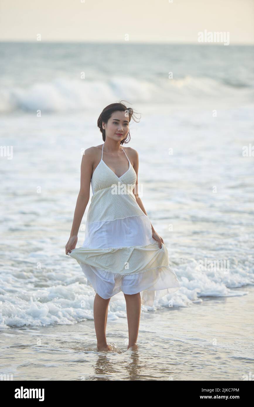 Portrait en longueur d'une femme asiatique attirante portant une robe d'été en regardant loin pensivement tout en se tenant debout à la cheville profondément dans l'eau de mer ondulée Banque D'Images