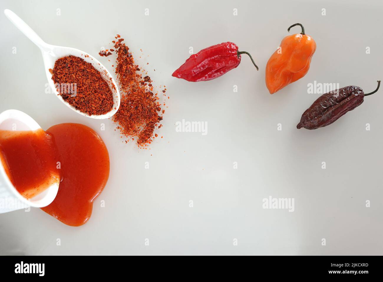 Trois variétés de poivrons chauds Scotch bonnet, naga morich, bhut jolokiaet leurs dérivés - sauce et poivre chaud moulu sur une surface en verre blanc Banque D'Images