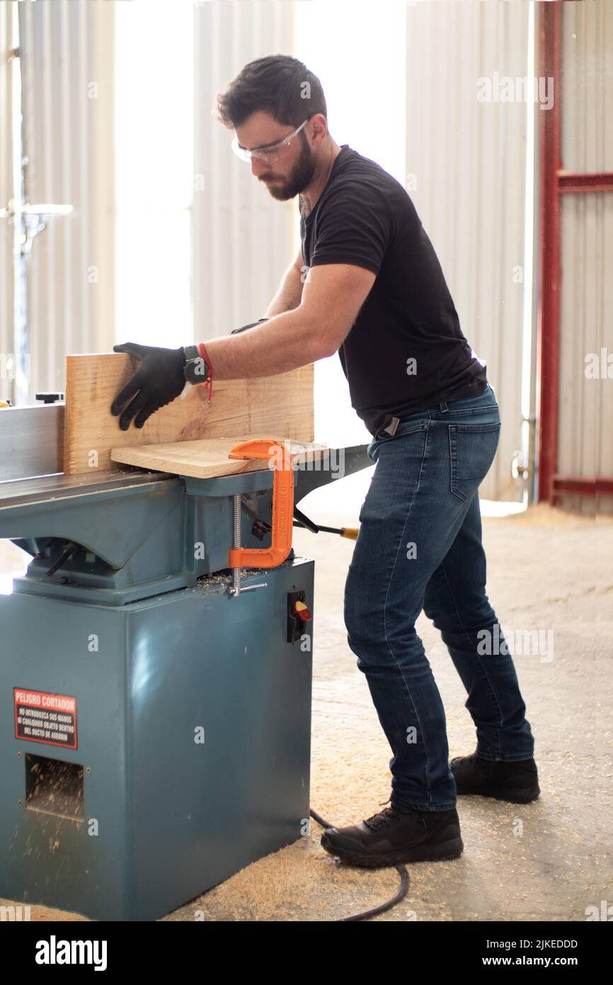 Mains d'un jeune menuisier travaillant sur une machine de jointer électrique de paillasse coupant une planche en bois dans un atelier de menuiserie à l'aide de gants noirs de sécurité Banque D'Images