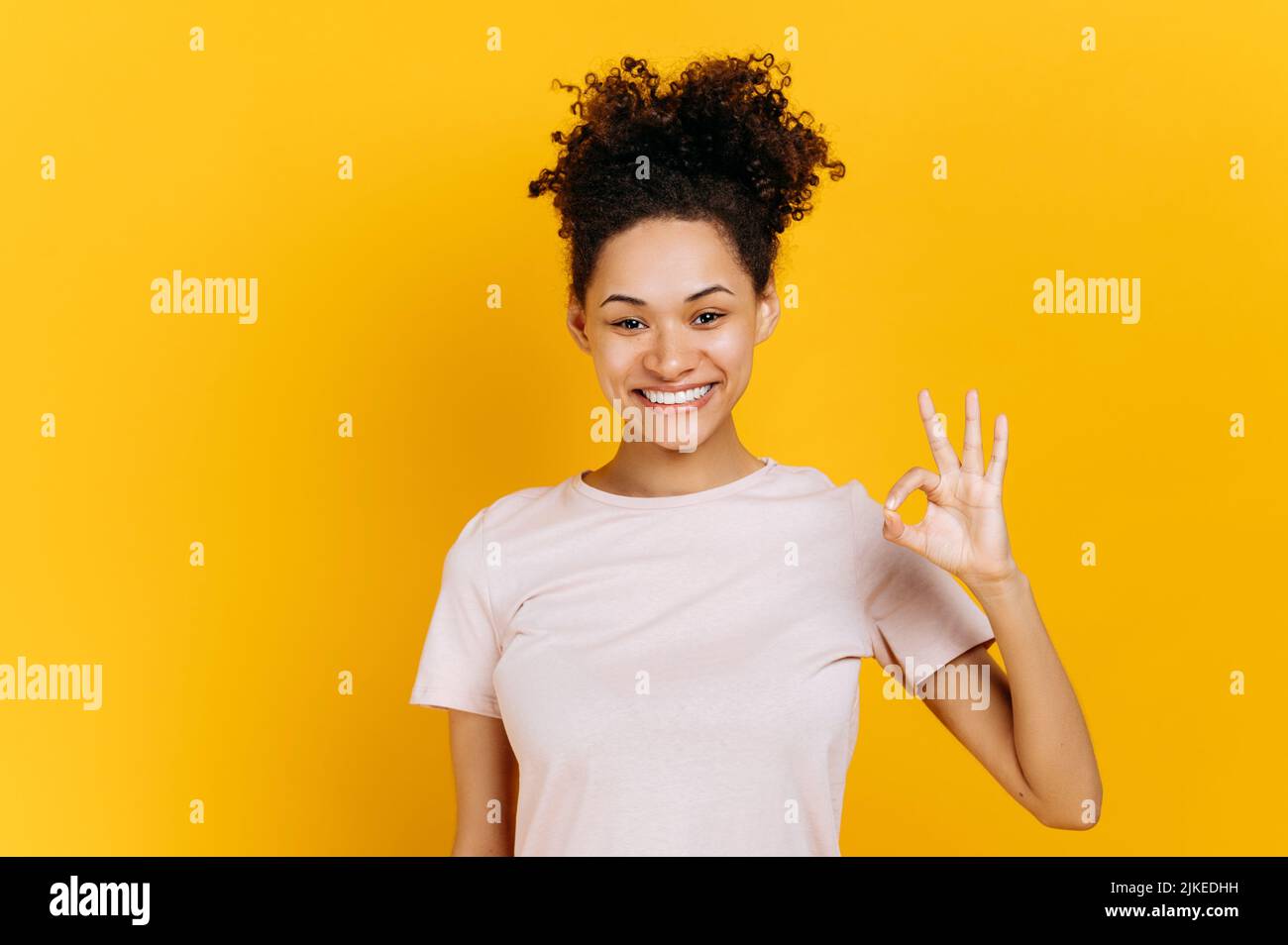 Positive heureuse confiante afro-américaine jeune femme à poil dur, fait avec le doigt OK symbole, montre signe d'accord, debout sur un arrière-plan orange isolé, regarde la caméra, souriant amical Banque D'Images