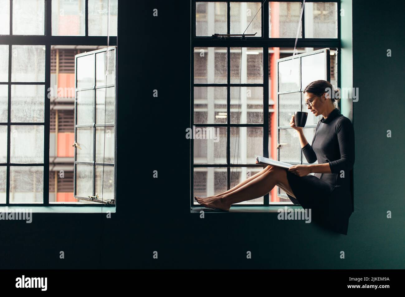 Femme asiatique assise sur le rebord de la fenêtre lisant un livre et buvant du café. Femme se détendant à l'intérieur avec un magazine et ayant un café. Banque D'Images