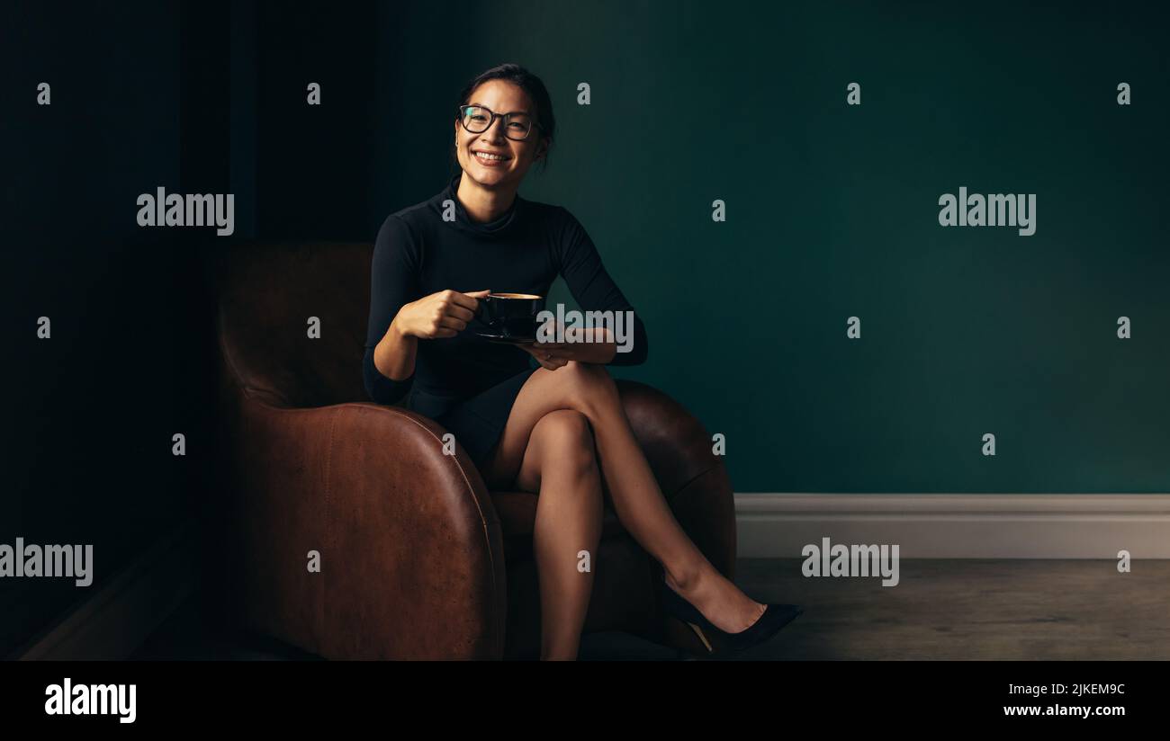 Femme souriante assise sur une chaise et tenant une tasse de café. Bonne femme asiatique en train de prendre un café dans un fauteuil. Banque D'Images