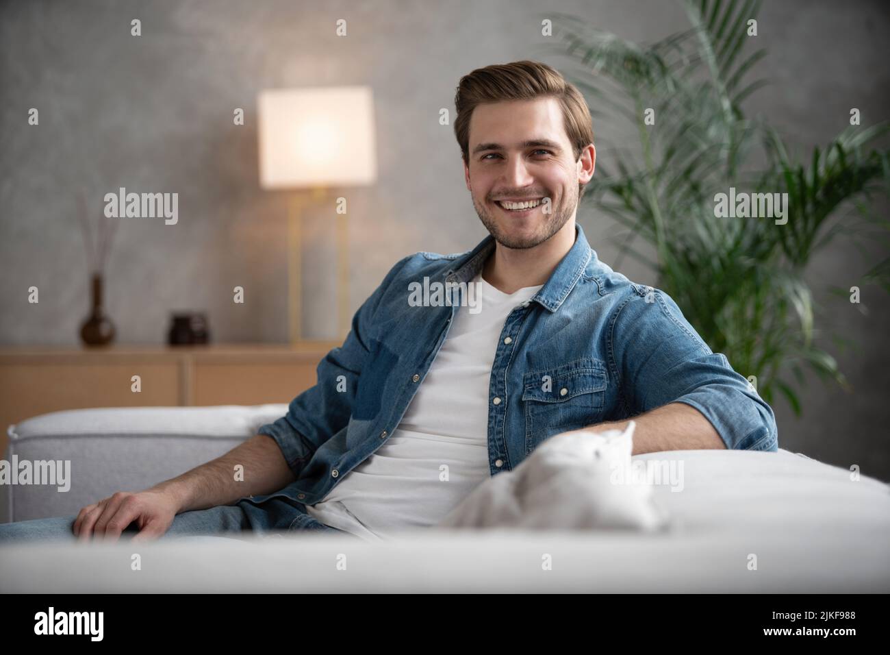 beau caucasien en chemise denim assis sur un canapé dans le salon regardant la caméra avec un sourire amical Banque D'Images