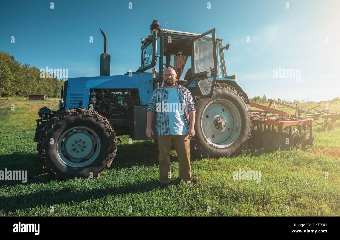 Portrait d'un fermier souriant et joyeux debout à l'arrière-plan du tracteur. Travailleur agricole et temps de récolte. Banque D'Images