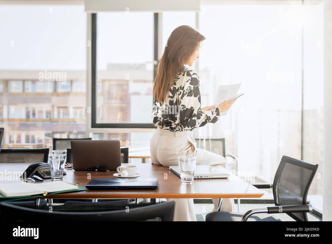 Femme d'affaires messagerie texte et tenue d'un document dans la main tout en étant assise dans un bureau moderne. Banque D'Images