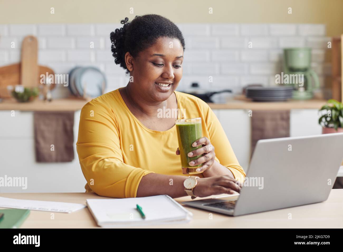 Portrait d'une femme noire souriante qui boit du smoothie et utilise un ordinateur portable à la maison Banque D'Images