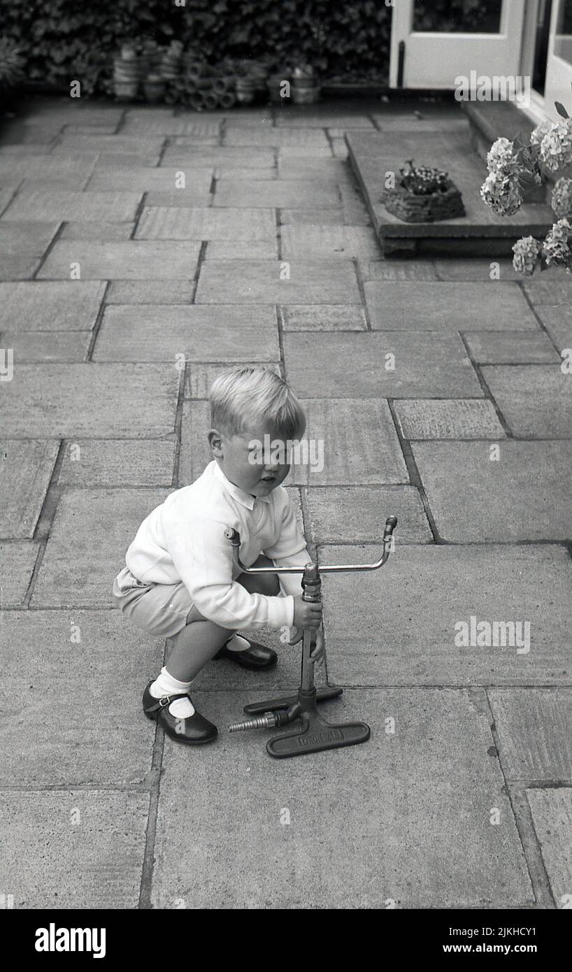 1960s, à l'extérieur sur un patio, un petit garçon heureux essayant de soulever un arroseur de jardin de l'époque, fabriqué par dron-Wal, en fonte et laiton, Angleterre. ROYAUME-UNI. Banque D'Images