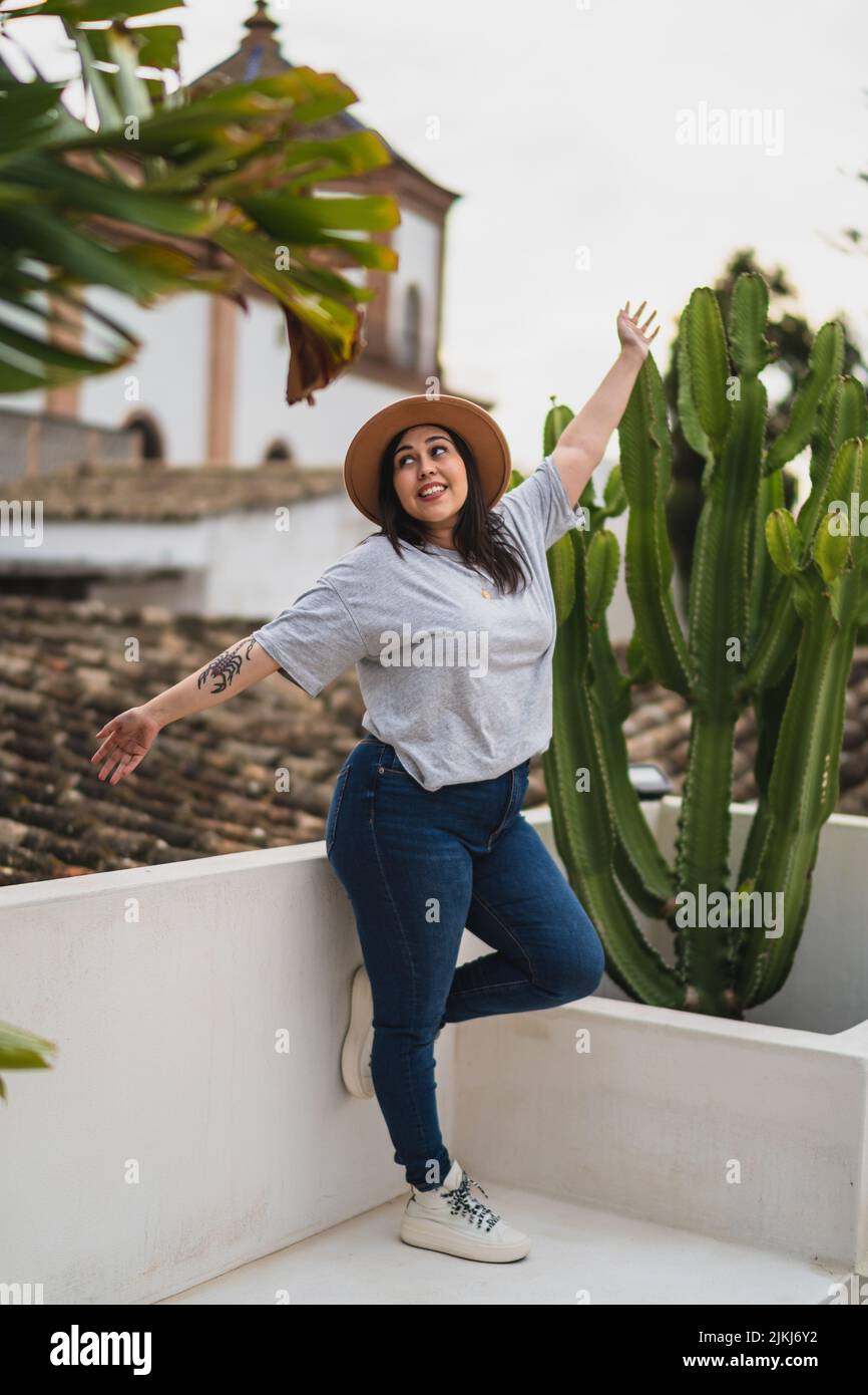 Une photo verticale d'une femelle caucasienne curvy avec un chapeau, souriant et posant contre un cactus Banque D'Images