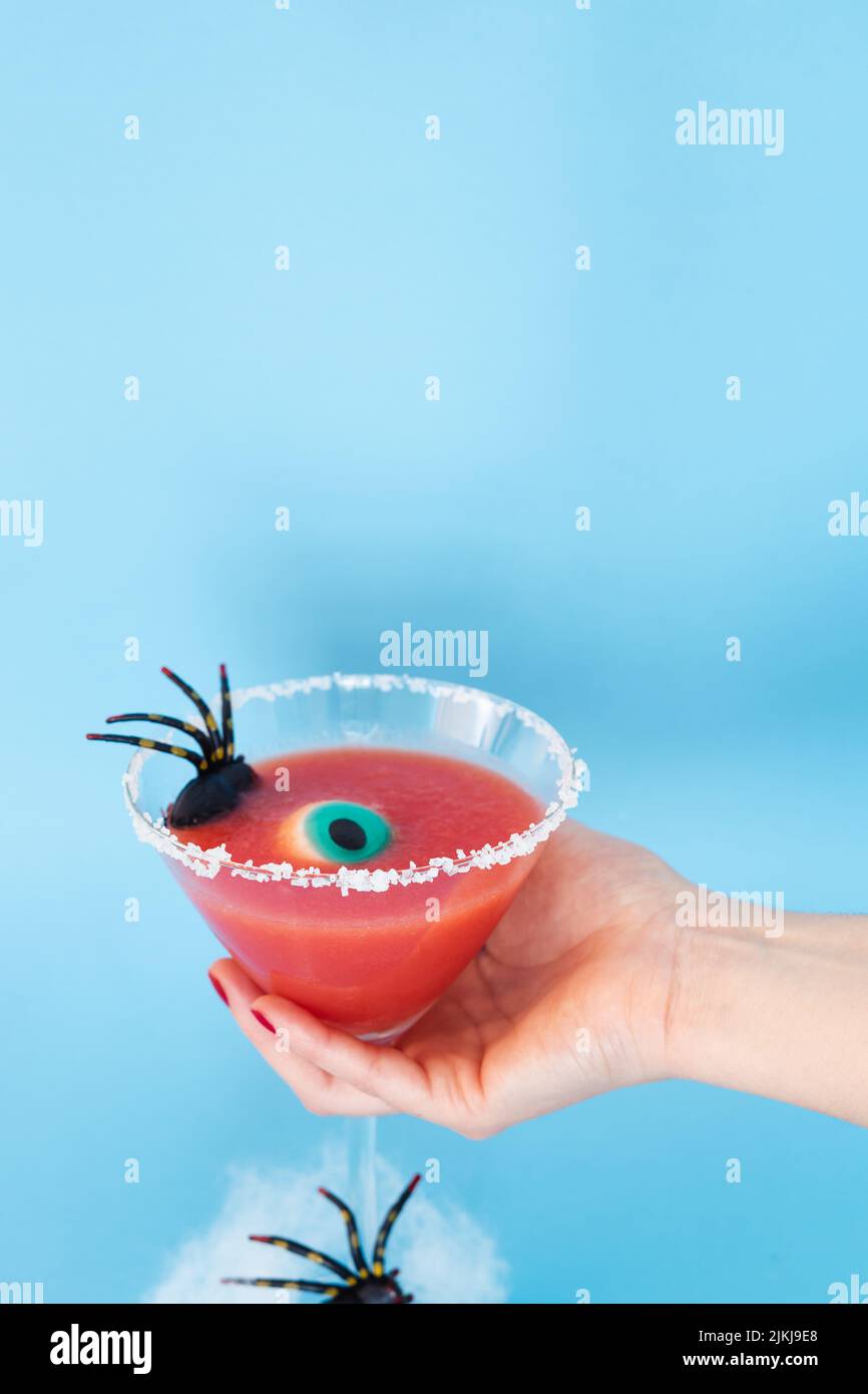 La main de femme tient un cocktail rouge effrayant avec la décoration d'Halloween sur fond bleu. Photo verticale. Banque D'Images