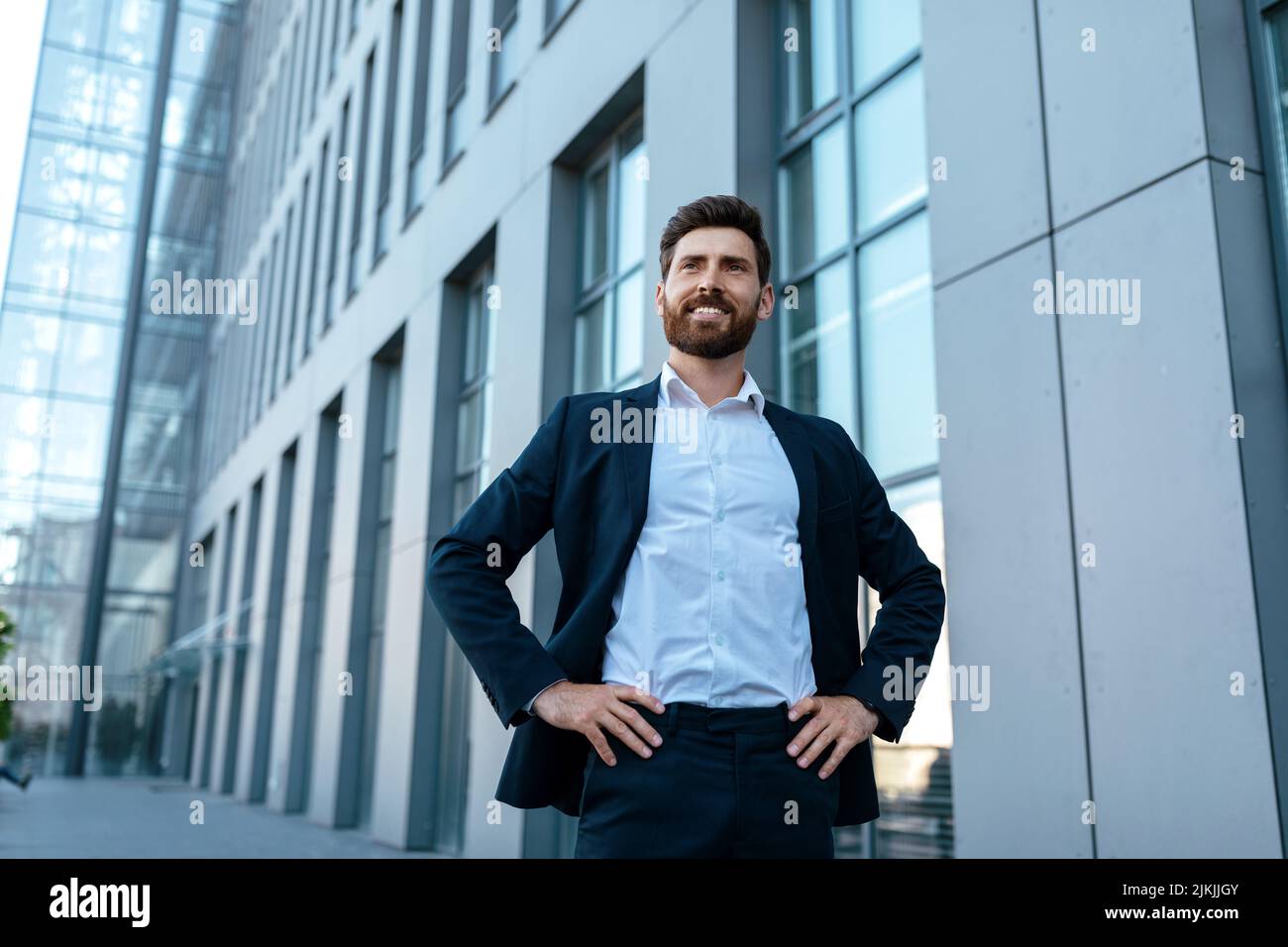 Joyeux homme d'affaires millénaire confiant et charmant avec une barbe en costume près d'un immeuble de bureaux moderne Banque D'Images