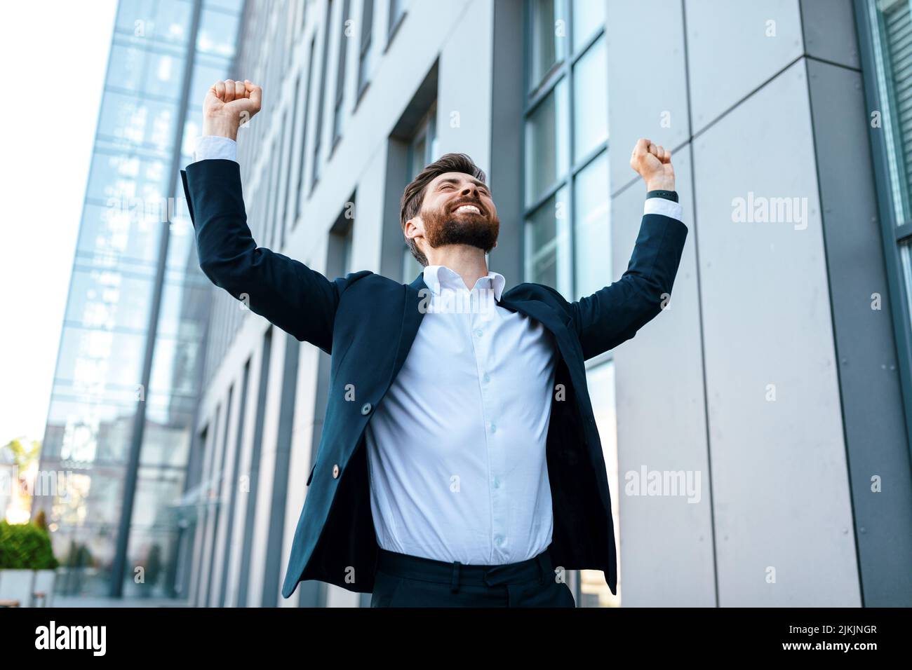 Joyeux et attrayant jeune homme d'affaires caucasien avec la barbe en costume lève les mains et se réjouit de la victoire Banque D'Images