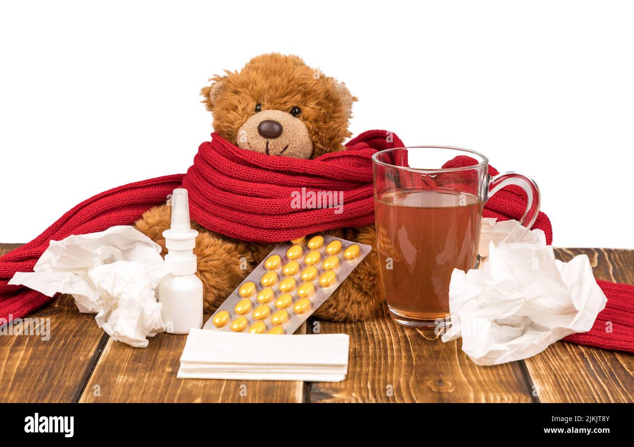 Un ours en peluche avec un spray nasal, des pilules, une écharpe rouge, du thé et des mouchoirs sur une surface en bois - souffrant de maladie Banque D'Images