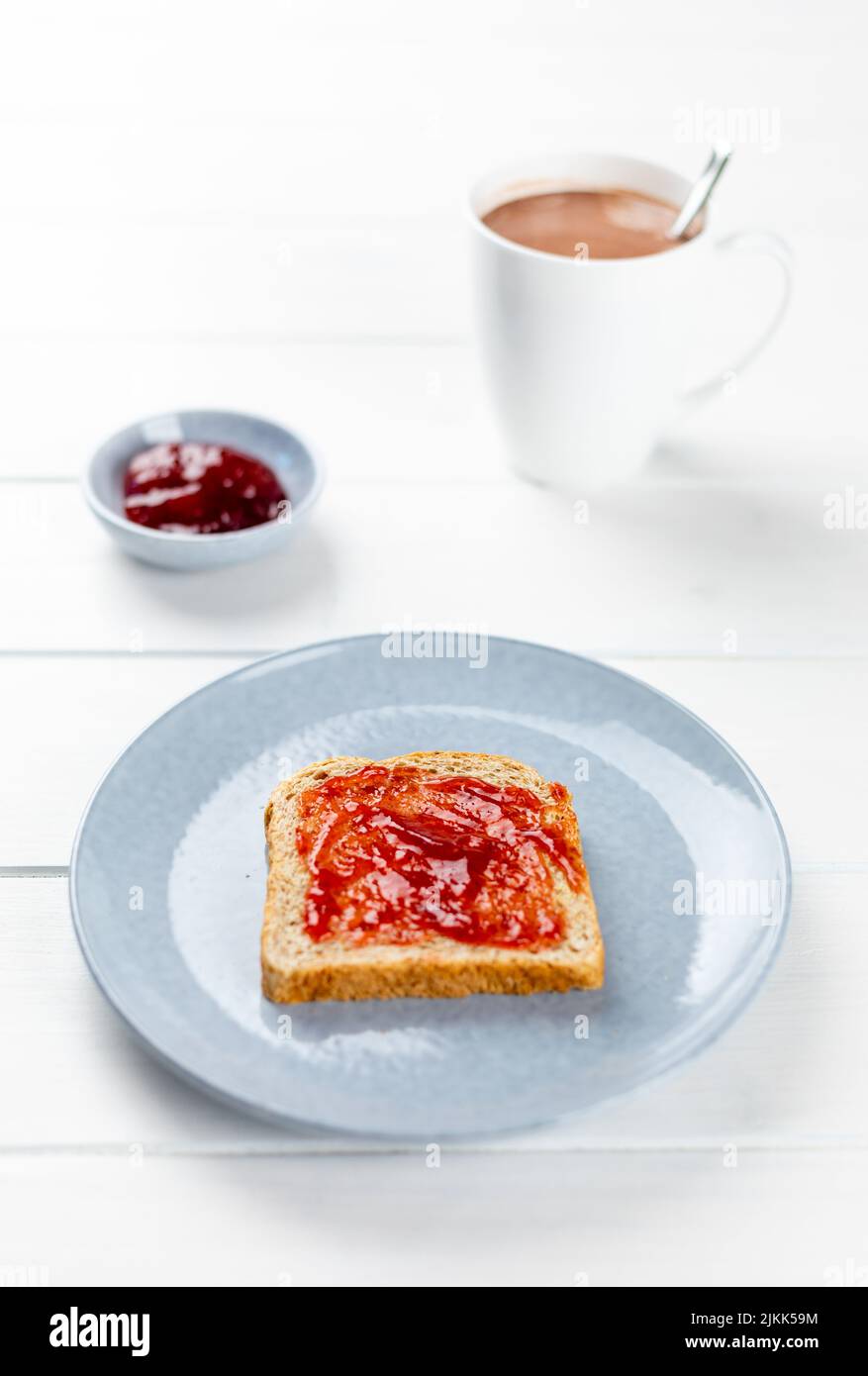 griller avec de la confiture de fraise sur une assiette bleue et une tasse de cacao sur fond blanc en bois Banque D'Images