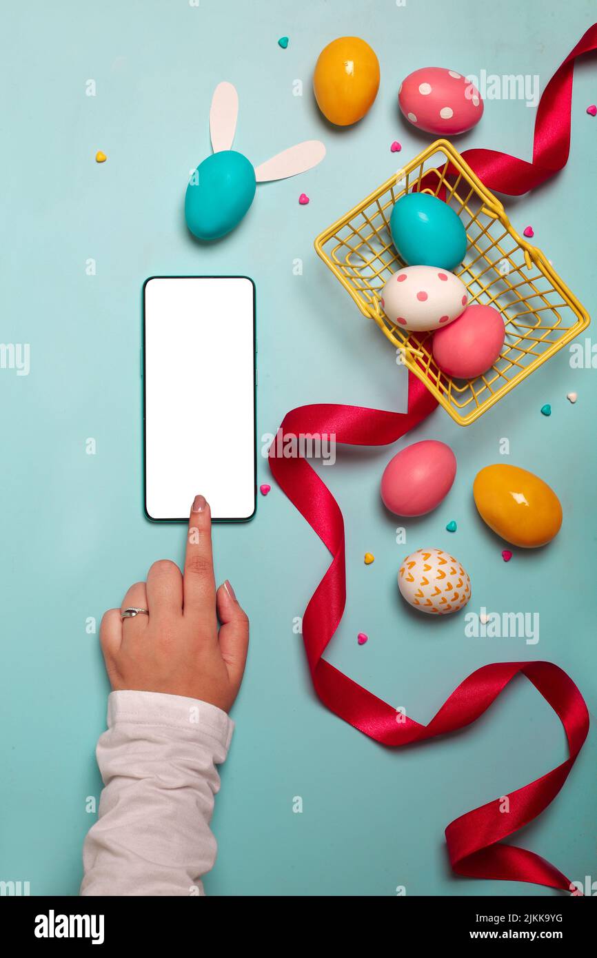 Vue de dessus d'un smartphone adulte avec des œufs de Pâques colorés, un ruban rouge et un petit panier jaune sur fond bleu clair Banque D'Images