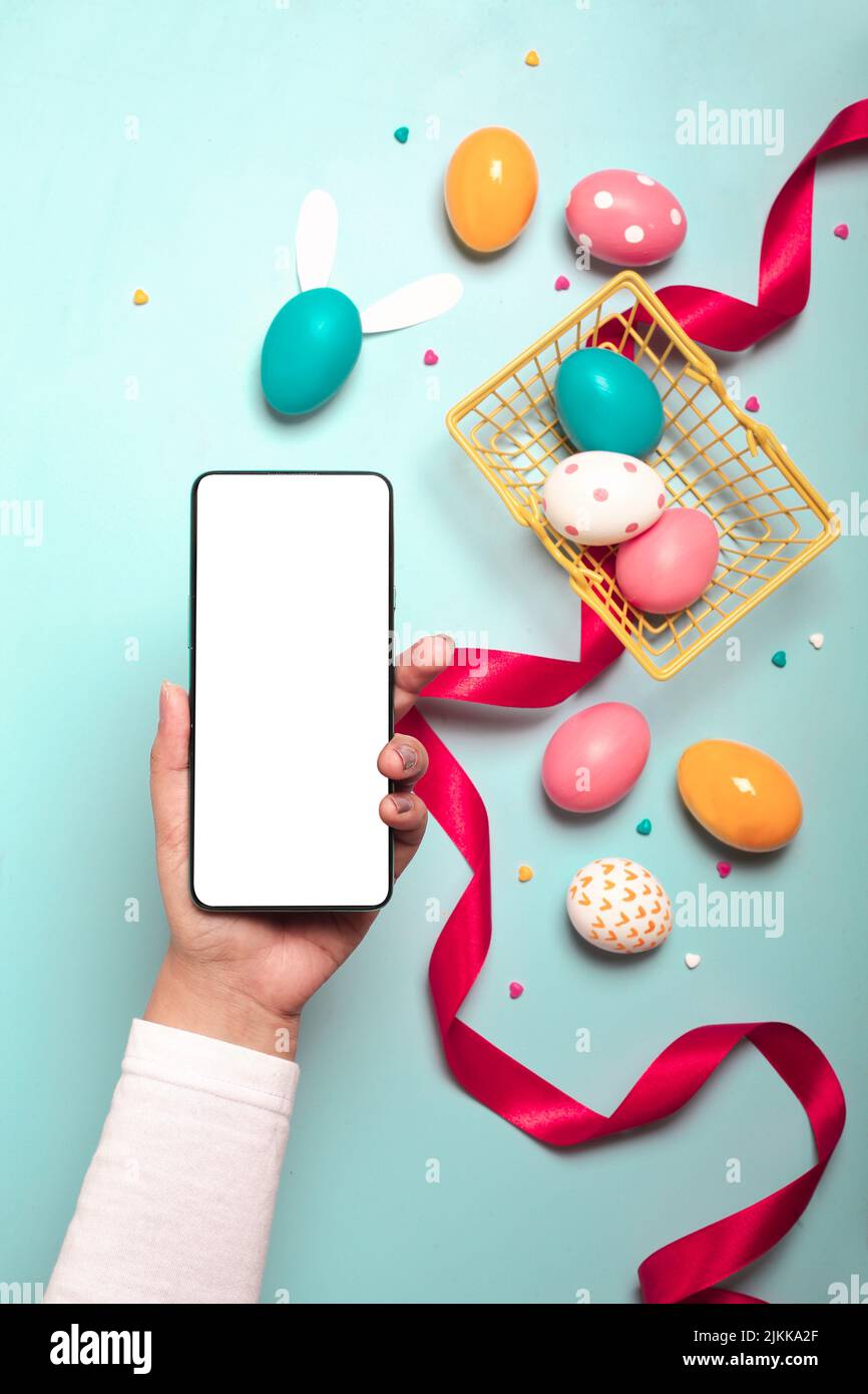 A top view of an adult hand holding smartphone with colorful Easter eggs, red ribbon and small yellow basket on a light blue background Banque D'Images