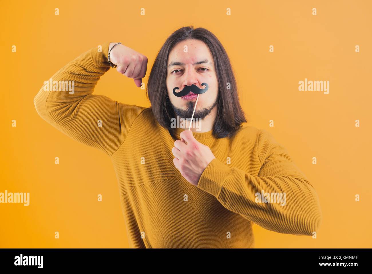 Beau garçon hispanique à la barbe, accroché à la moustache en papier et aux muscles serrés sur fond jaune dans un studio. Photo de haute qualité Banque D'Images