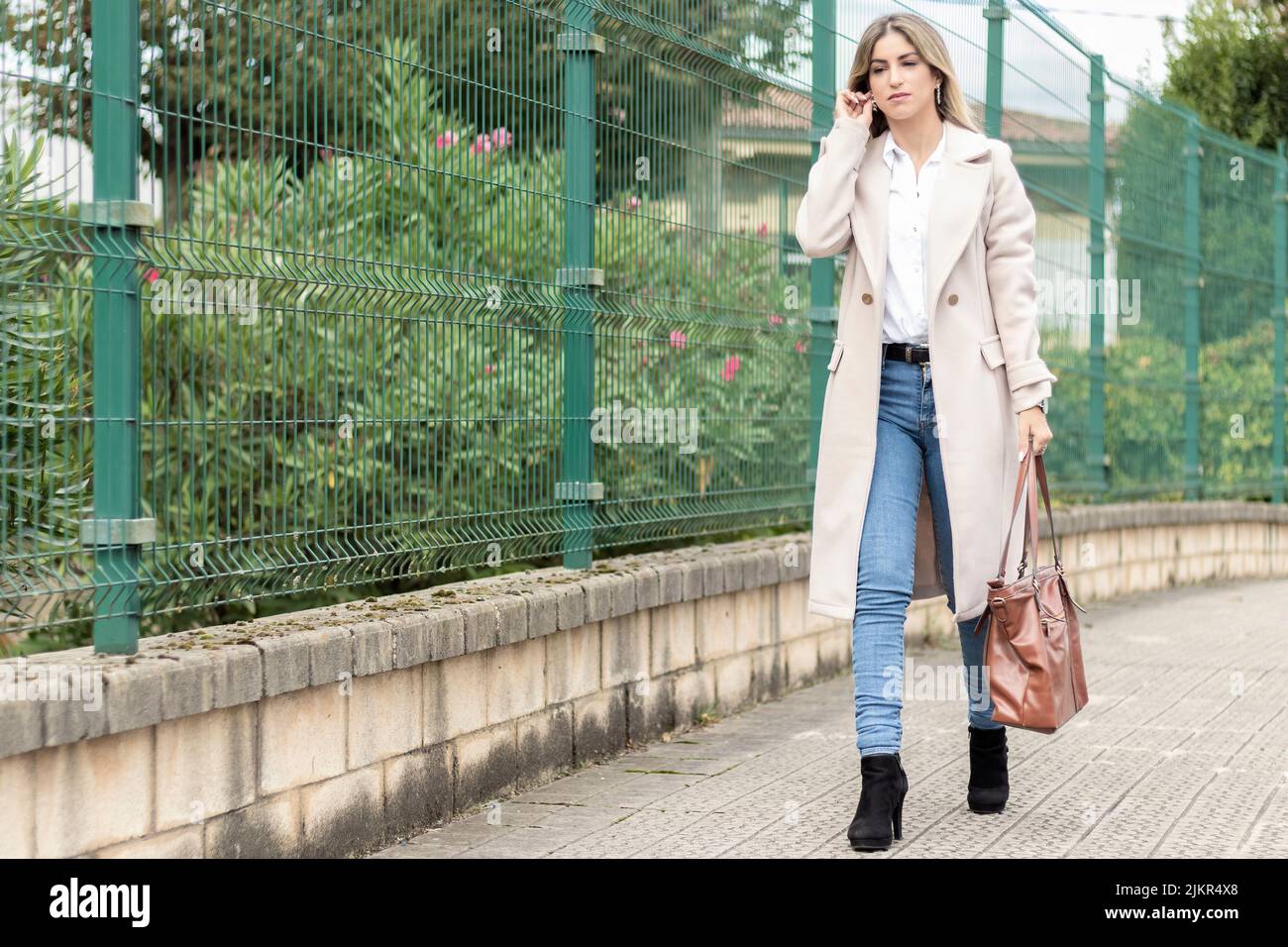 femme d'affaires marchant dans la rue avec un sac marron un manteau beige, une chemise blanche et un jean Banque D'Images