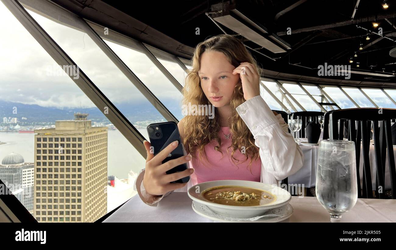 Une adolescente dans une chemise blanche aux cheveux blonds s'assoit et regarde le téléphone iPhone 13 qu'elle est intéressée et avec son doigt quelque chose corrige le téléphone noir Top of Vancouver Revolving Restaurant. Banque D'Images