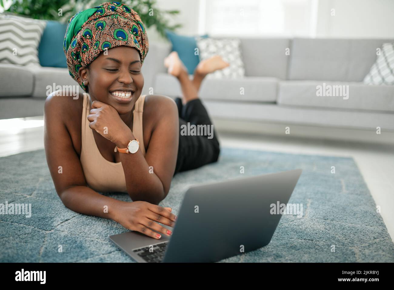 Souriante jeune africaine Am assis avec ses jambes croisées sur son tapis à la maison à l'aide d'un ordinateur portable et regardant dans l'appareil photo Banque D'Images