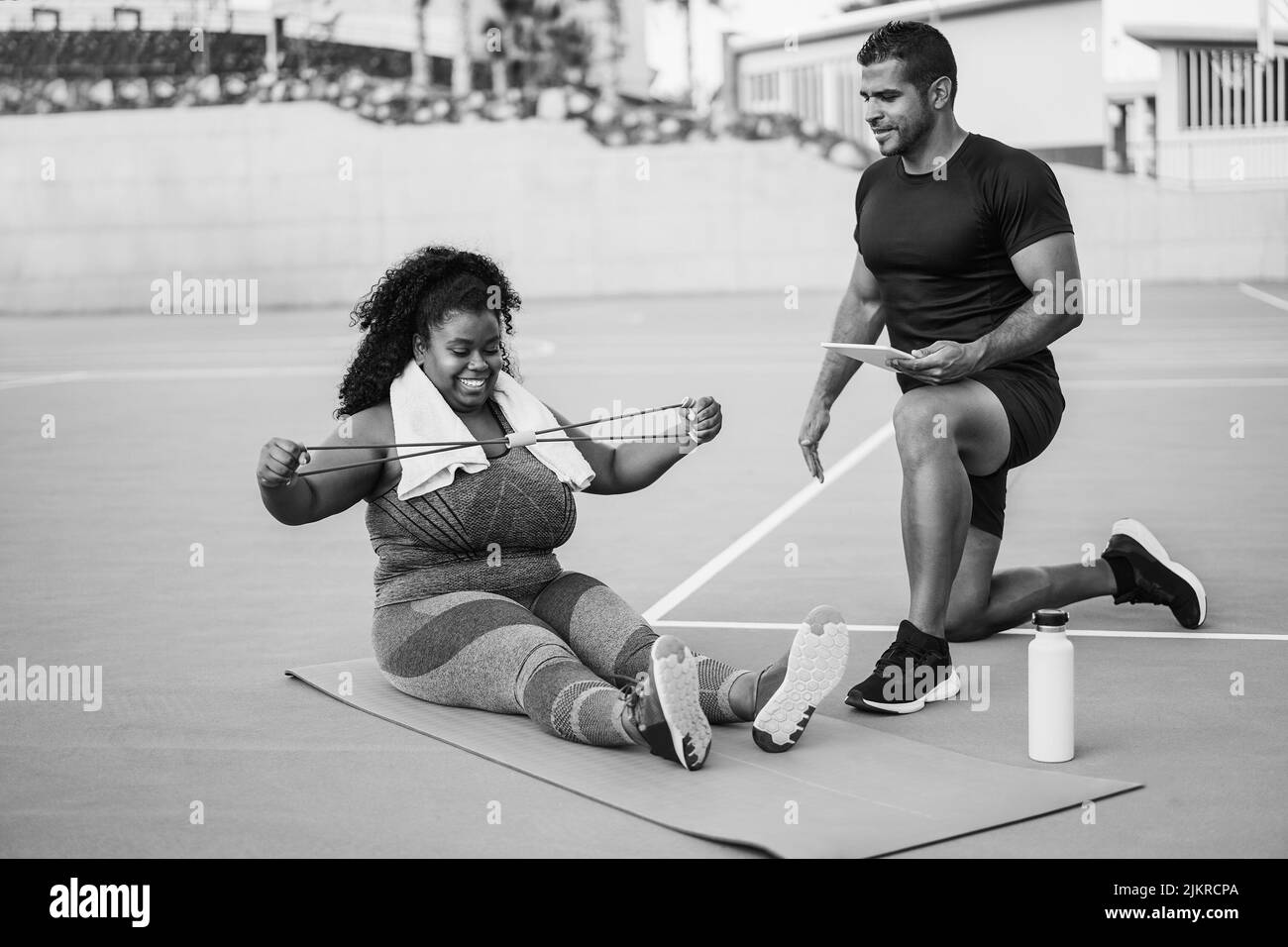 Femme curvy et entraîneur personnel faisant la séance d'entraînement en extérieur - Focus sur les mains de fille tenant la bande d'étirement - montage noir et blanc Banque D'Images