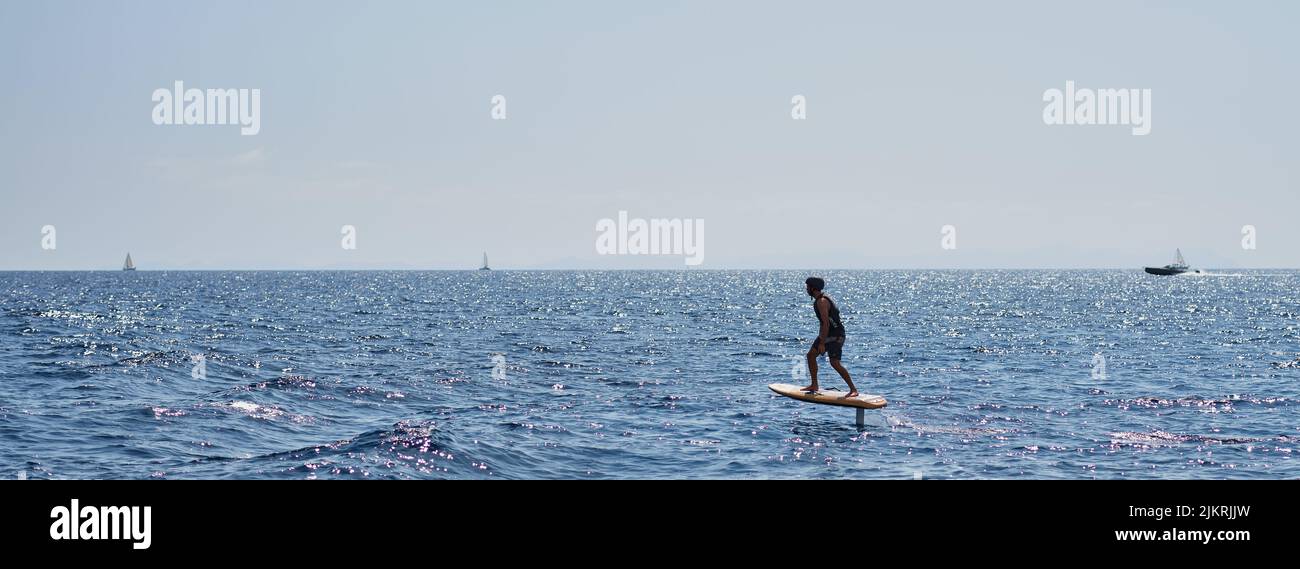 Cavalier sur une planche de surf électrique sur la mer calme avec des voiliers lointains à l'horizon dans un large paysage marin panoramique Banque D'Images