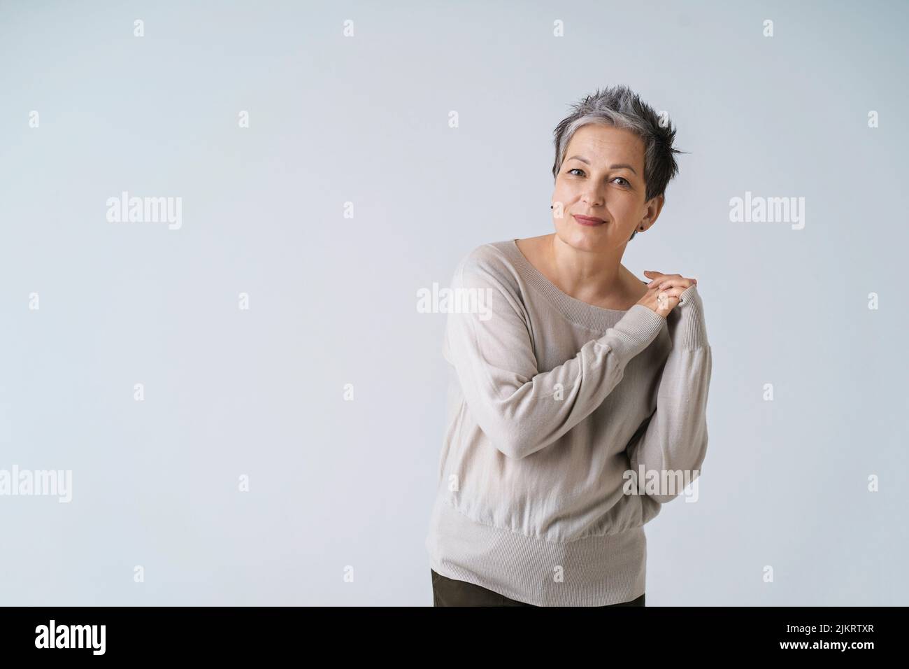 Souriante mature gris cheveux courts femme posant avec les mains repliées sur son épaule regardant l'appareil photo portant un chemisier blanc, espace de copie isolé sur fond blanc. Soins de santé, concept de beauté vieilli. Banque D'Images