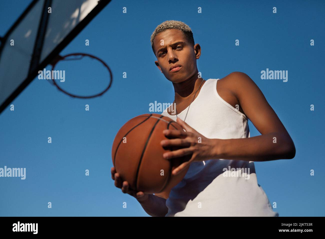 De dessous de sérieux joueur de basket-ball américain africain avec le ballon regardant la caméra près de la boucle contre le ciel bleu sur le terrain de jeu Banque D'Images