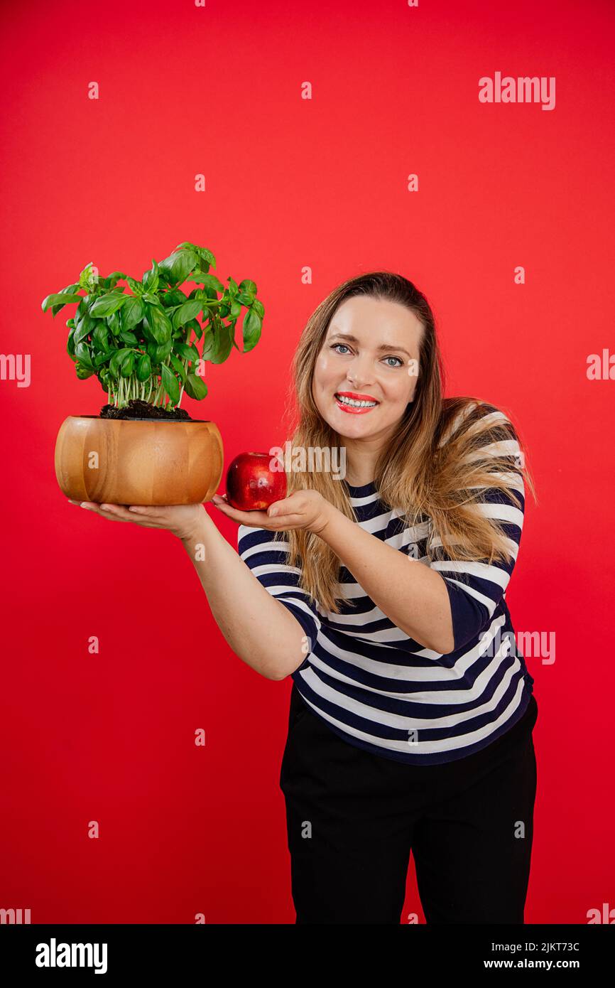 Vertical souriant Plump femme en chemise rayée regarder l'appareil photo tenant le pot de plante de basilic vert et la pomme juteuse. Nouvelle recette Banque D'Images