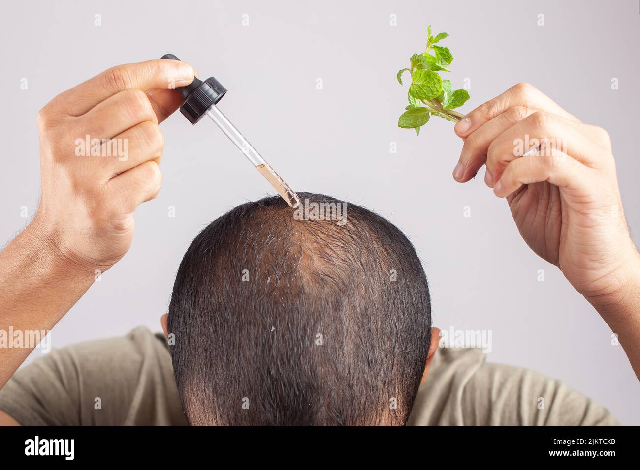 jeune homme montrant des feuilles de menthe et appliquer l'huile essentielle de cheveux dans les taches chauves sur fond gris. Banque D'Images