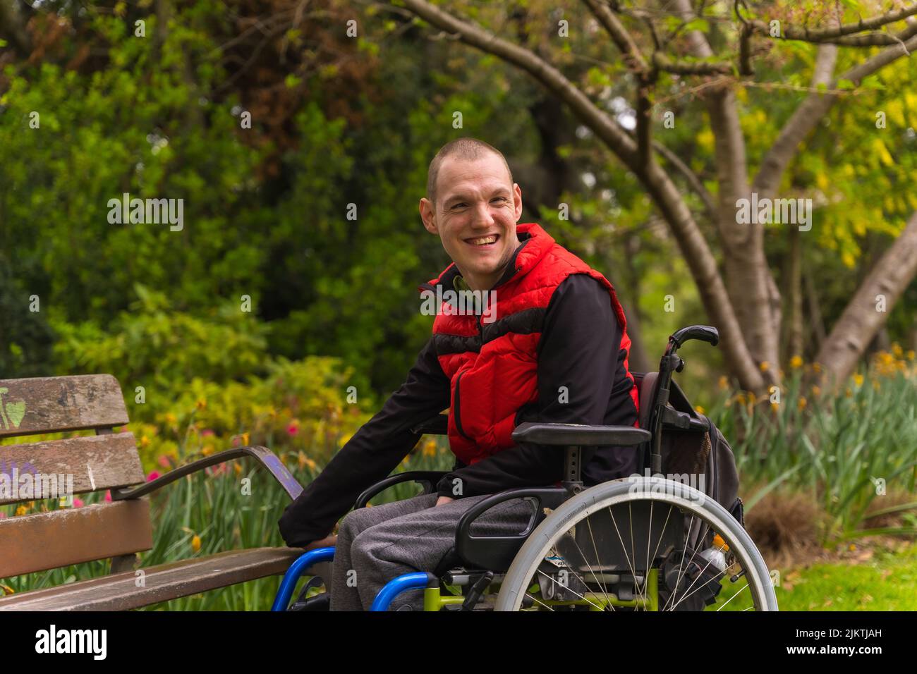 Portrait d'un jeune homme paralysé dans un gilet rouge dans un parc public de la ville. Assis en fauteuil roulant à côté d'une chaise de parc Banque D'Images